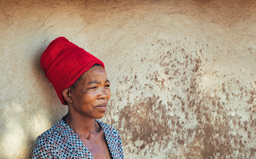 old African woman with red scarf, natural light against the wall of the mud house in the yard of her house in the village