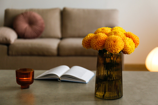 Beautiful bouquet of yellow dahlia flowers in a glass vase, lit candle and an open book on a grunged stone tabletop. Interior background, close up, copy space.