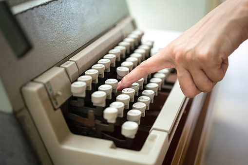 Action of a human's hand is typing on a traditional typewriter which is placed on working desk. Equipment object photo. Close-up and selective focus.