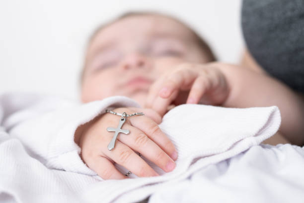 close-up detail of a baby's hand at his baptism, holding a christian cross. young mother holding her baby at her catholic baptism in latin church. caucasian baby with a silver cross in his hand. close-up detail of a baby's hand at his baptism, holding a christian cross. young mother holding her baby at her catholic baptism in latin church. caucasian baby with a silver cross in his hand. god is love stock pictures, royalty-free photos & images