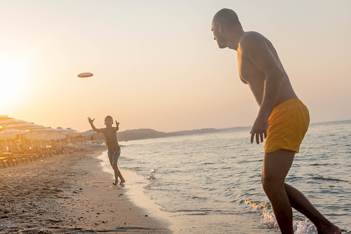 Father and son playing frisbee together in shorts at sea sunset