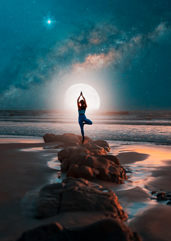 silhouette of a woman in yoga pose on rocks on the beach at night with the moon and milky way in the background