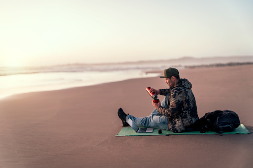 mature man backpacker or digital nomad sitting on the shore of the beach with laptop and drinking coffee or tea