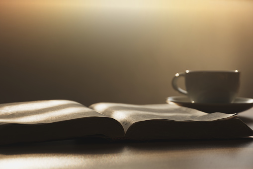 Open the Bible in the morning for prayer with a cup of coffee on a wooden table.  with the morning sun shining through the window