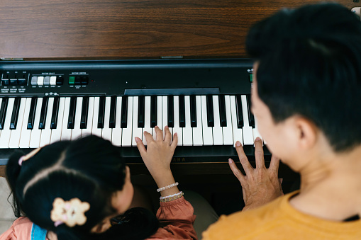 Cheerful Asian daughter in casual clothes with pigtails smiling and looking at dad while leaning to play piano in daytime at home