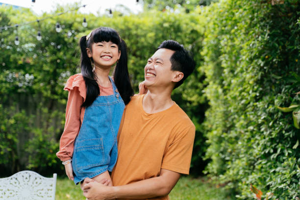 glücklicher asiatischer vater in freizeitkleidung, der eine kleine tochter trägt und mit einem lächeln in die kamera schaut, während er tagsüber im hinterhof in der nähe von büschen steht - offspring child toothy smile beautiful stock-fotos und bilder