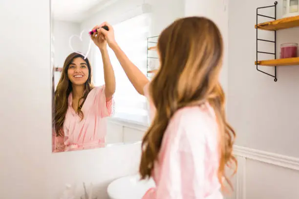 Happy latin woman seen from behind drawing a heart in the bathroom mirror with a lipstick during a self care day