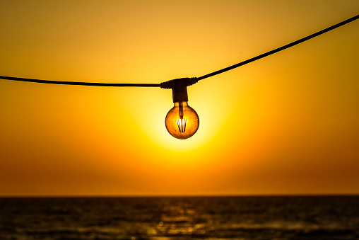 Concept of solar energy. Sun shines through the modern energy saving filament led bulb edison style light lamp against of sky and sea in the sunset. Ecology concept.