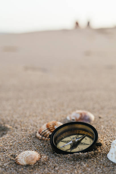 istock vertical image for mobile devices of a compass next to seashells on the wet sand on the shore in front of the ocean. in the background two silhouettes of a couple walking during summer vacation. 1411661572