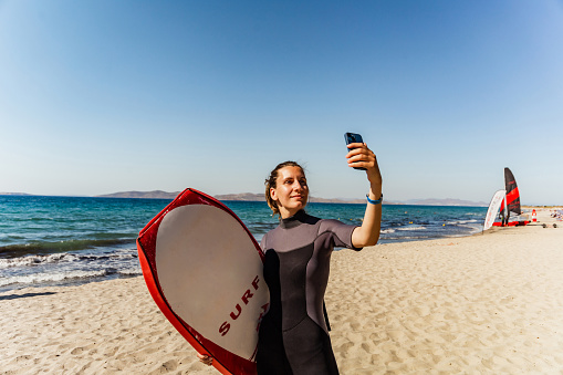 Portrait of a young female surfer holding a surfboard and taking a selfie on a beautiful sandy beach
