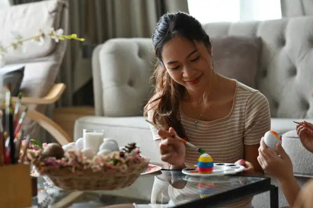 Smiling Asian girl sit on ter floor while painting multi-color Easter egg on the colorpalette on coffee table with her family.