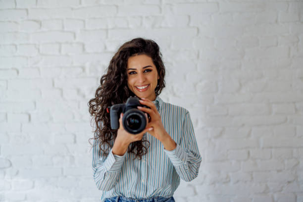 You look great in this picture A young Caucasian woman with a bright smile is standing in front of a wall and cheerfully looking at the photographer while taking a picture with her camera. Photographer stock pictures, royalty-free photos & images