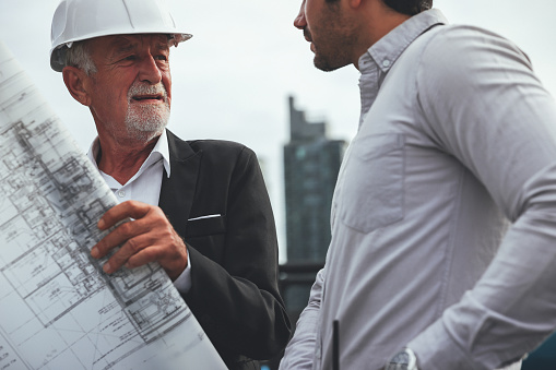 An architect or civil engineer with planning documents meets with a customer or colleague on the large construction site and both of them wear white protective helmets. Foundation under construction can be seen in the background. Photographed in high resolution with copy space