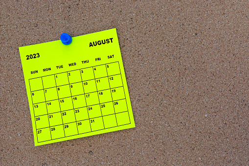 August 2023 yellow sticky note calendar with pin on cork bulletin billboard. Copy space.
