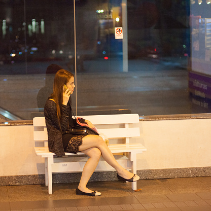 Thai woman is talking at mobile phone and is sitting on a bench at night. Scene is outside of MRT station at Ladprao Road in Bangkok. Woman is wearing a skirt and has legs crossed