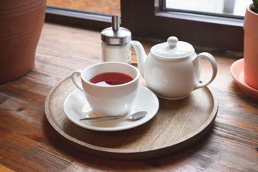Cup with red fruit tea, teapot and sugar bottle on round wooden tray on wooden window sill, cafe. Front view.