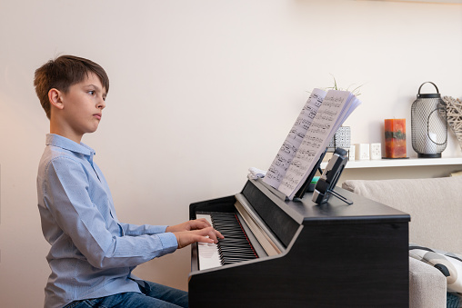 A brunette tidy boy in a blue shirt at home in a bright room contented and focused plays the electric piano to the notes of the composer. The boy is happy, sad, giving signs of success, thumbs up, listening to the piano with headphones, wearing a headband, helping himself with a phone app. Taking selfie