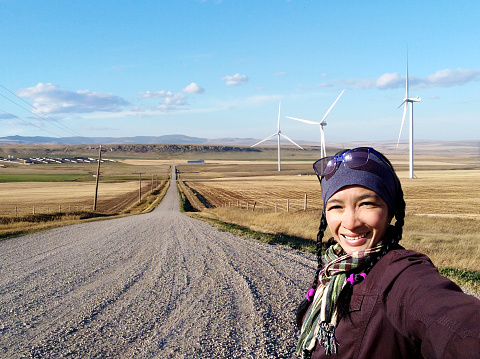 A young woman person taking a selfie with a wind turbine and gravel country road background. Travel destinations in Pincher Creek, Canadian Prairies in Alberta, Canada.