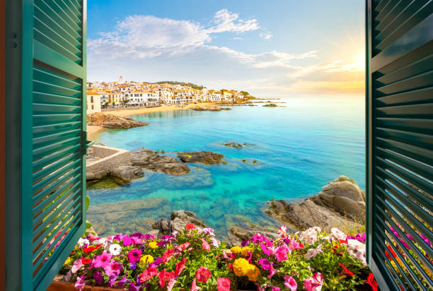 blick durch ein offenes fenster mit fensterläden auf den sandstrand, die felsige küste und die weiß getünchte stadt calella de palafrugell, spanien, an der küste der costa brava, wenn die sonne untergeht. - strandurlaub stock-fotos und bilder