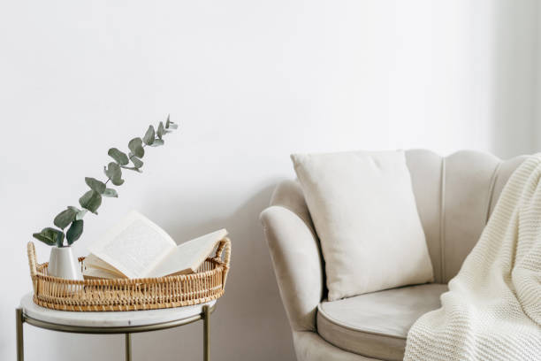 Open book on tray in room, near beige armchair with pillow Wicker tray and open book close to eucalyptus branch in ceramic vase on side table. Bohemian design of living room with beige soft chair with cushion and white plaid bamboo plant photos stock pictures, royalty-free photos & images