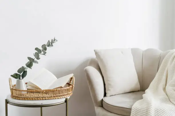 Wicker tray and open book close to eucalyptus branch in ceramic vase on side table. Bohemian design of living room with beige soft chair with cushion and white plaid