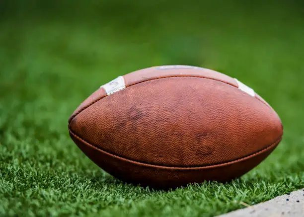 A pigskin leather football lies on the sidelines during a college game. Graphics removed from ball.