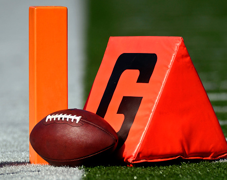 An American pigskin  football and goal markers lie in on the artificial turf prior to an NFL game. Note: Graphics altered from football.