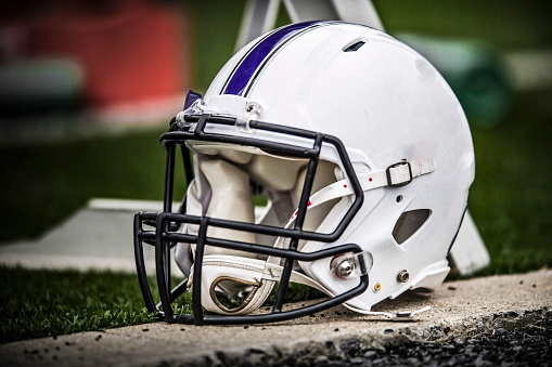A White Football Helmet with purple stripe lies on the sidelines during an American college football game. Note: Graphics removed from helmet.