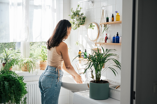 Young beautiful woman with dark skin washing hands in light bathroom with many green plants. Biophilic design of interior. Natural cosmetics, self care and wellness concept