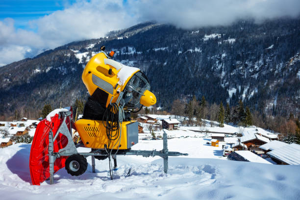 Yellow snow canon at ski resort with mountains on background on sunny day, snow-gun spraying artificial ice crystals.