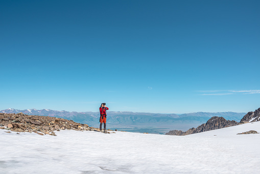 Tourist in red walks on snow mountain near abyss edge on high altitude under blue sky in sunny day. Man with camera on snowy mountain near precipice edge with view to large mountain range in away.