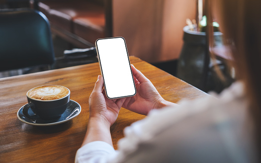 Mockup image of hands holding mobile phone with blank desktop screen with coffee cup on wooden table
