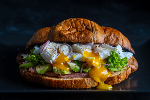 Croissant fresh sandwich with poached egg, salmon and avocado on a plate on black background, close up