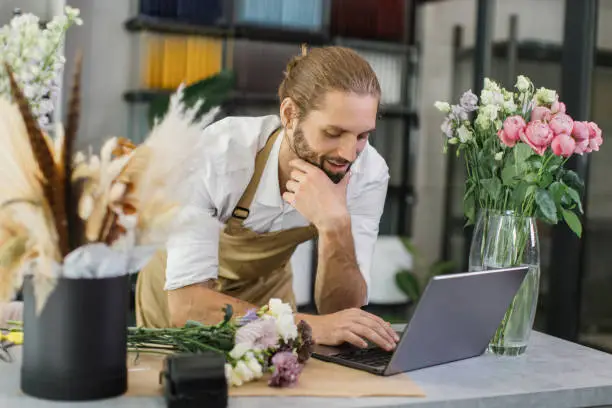 Bearded man florist discusses the order by talking on video call, using laptop in a flower shop, working online, male chooses flowers for the customer.