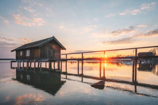 A wooden pier extends into the calm waters of Chiemsee during sunset in Bavaria, Germany. The sun reflects on the lake’s surface, creating a tranquil and atmospheric scene under a clear evening sky.