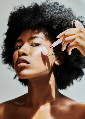African woman using stone roller to massage her face against studio background. Beautiful afro American model giving herself a facial treatment with a cosmetic massaging tool on her glowing skin