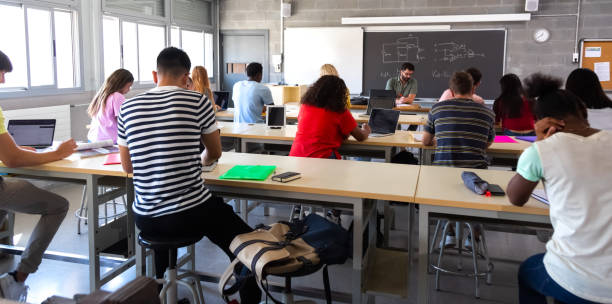 vista posteriore di un gruppo di studenti delle scuole superiori multirazziali in classe che utilizzano laptop mentre l'insegnante segna gli esami. - grading teacher desk writing foto e immagini stock