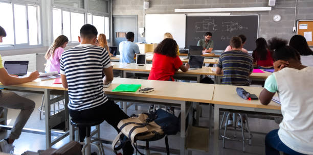 rear view of group of multiracial high school students in class using laptops while teacher marks exams. - college student high school student education learning imagens e fotografias de stock