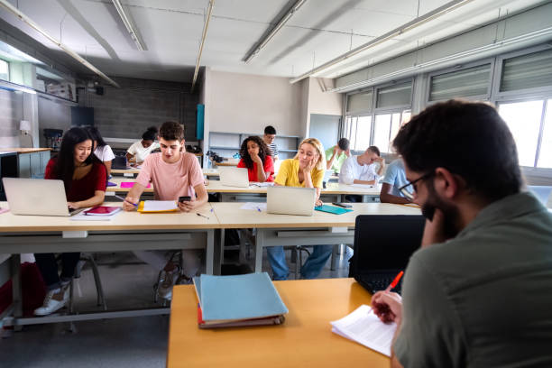 enseignant caucasien masculin marquant les examens tandis que les étudiants multiraciaux étudient et utilisent des ordinateurs portables en classe. - grading photos et images de collection