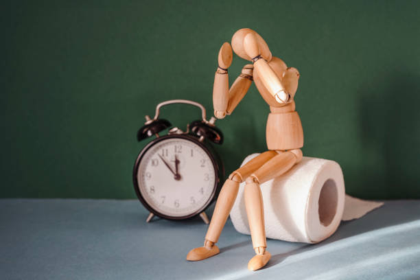 Wooden figure sit on a roll of toilet paper near alarm clock. Concept of the problem with digestion. stock photo
