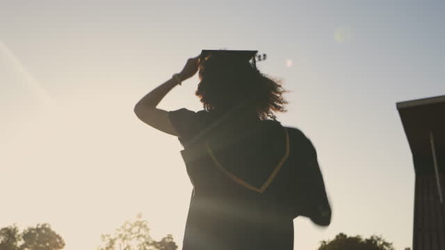 A female student celebrating her graduation by cheering and raising her arms outside. Rearview silhouette of a graduate wearing a cap and hat, feeling excited after her graduation ceremony at sunset