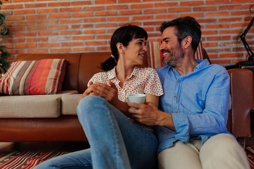Middle-aged couple drinking coffee and looking at each other, sitting on the floor in front of the sofa