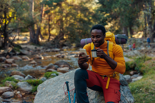 Young African ethnicity man video chatting via cellphone with somebody and drinking hot beverage as he having a hiking walk in the forest. Happy people, network technology, or traveling concept