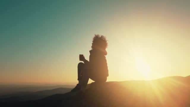 Rear view of woman hiker sitting on rock on top of hill while looking at sunset over mountain valley, relaxing drinking energy drink