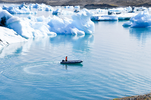 Man driving a dinghy between some glacier icebergs moving towards the sea with snow-capped mountains in the background in Iceland Europe