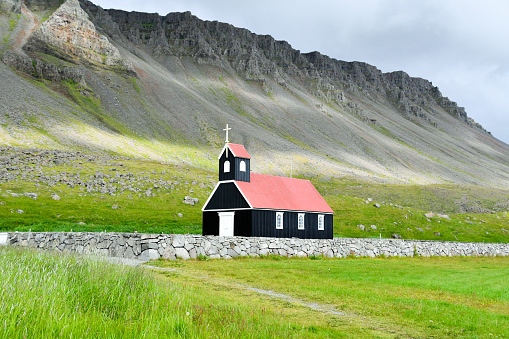 Small church with black walls and red roof in the middle of a meadow and the mountains illuminated by rays of sun behind Northen Europe