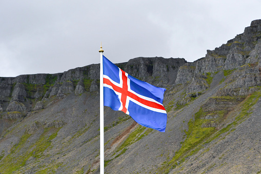 Blue flag with red and white cross of icelandic island fluttering with mountains as background