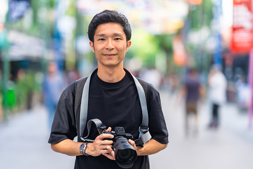 A portrait of a young male tourist with his camera looking at the camera and smiling in the street.