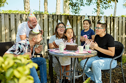 Mature gay male couple, lesbian moms, and sister watching, smiling, and applauding in support of 5 year old girl’s outdoor birthday celebration.