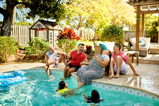 Mature gay male and lesbian couples sitting with legs dangling in water, talking, and laughing while young girls play and swim.